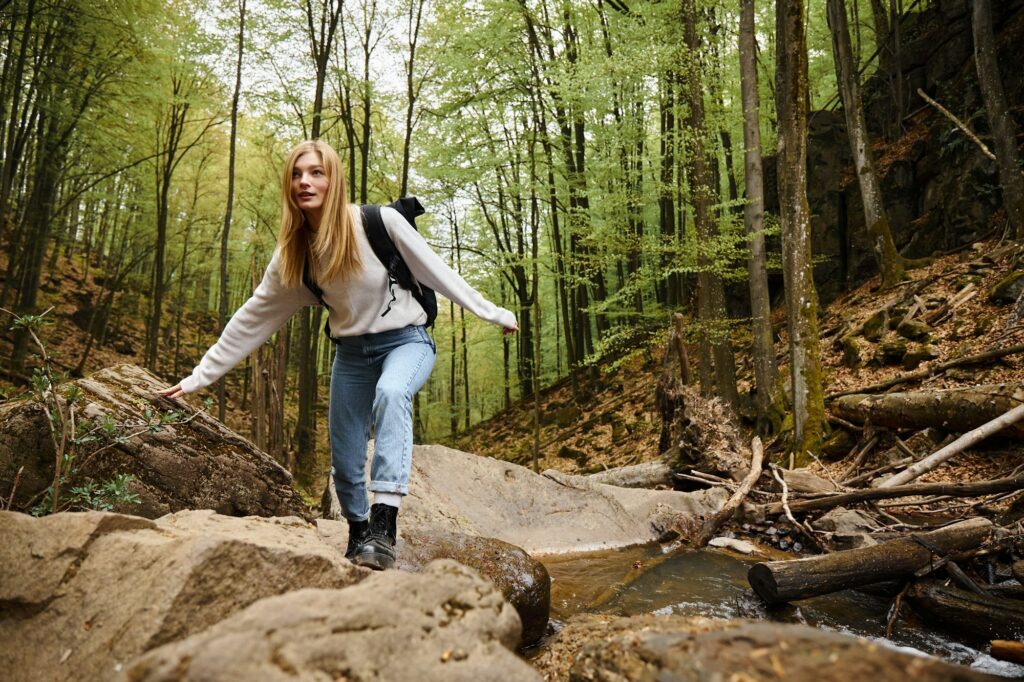 Woman with backpack hiking and balancing on mountain river rock in forest. Solo female tourist